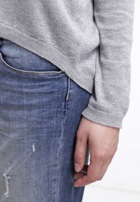 Hellgrauer Strickpullover mit abgerundetem Saum, kombiniert mit blauen Denim-Jeans, die Verwaschungen aufweisen und eine taillierte Silhouette haben.