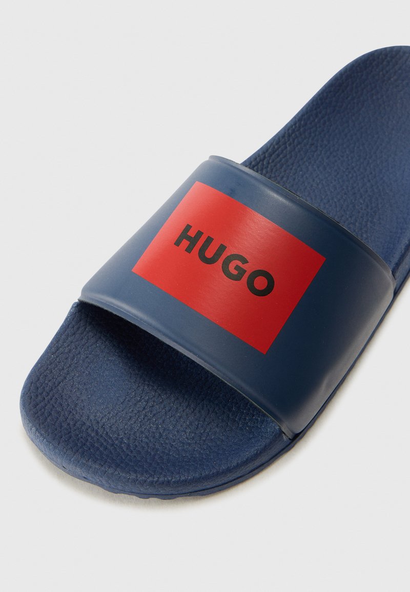 Granatowe klapki wykonane z gumy z teksturowaną podeszwą. Posiadają szeroki pasek z czerwonym prostokątem i czarnym napisem "HUGO."