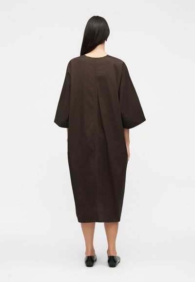 Γυναίκα με μακριά σκούρα μαλλιά, που φοράει ένα φαρδύ, σκουρό καφέ φόρεμα μέχρι το γόνατο με μανίκια τριών τετάρτων, στέκεται με την πλάτη προς τον θεατή.