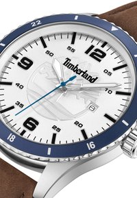 Reloj plateado con esfera blanca, bisel azul, correa de cuero marrón, marcadores de hora negros y segundero azul. Presenta el logo de Timberland.