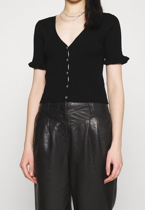 Femme portant un cardigan noir côtelé à manches courtes avec des boutons, des poignets volantés, et un pantalon en cuir noir taille haute.