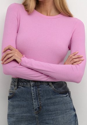 Vrouw met gekruiste armen, gekleed in een roze geribbelde top met lange mouwen en een blauw denim spijkerbroek met hoge taille, tegen een effen achtergrond.