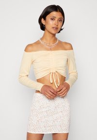 Top cropped em tom bege claro, com detalhes franzidos e laço na frente; combinado com uma saia mini branca com padrão floral.