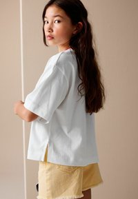 White short-sleeve t-shirt with side slits, paired with light yellow denim shorts featuring frayed edges and back pockets.