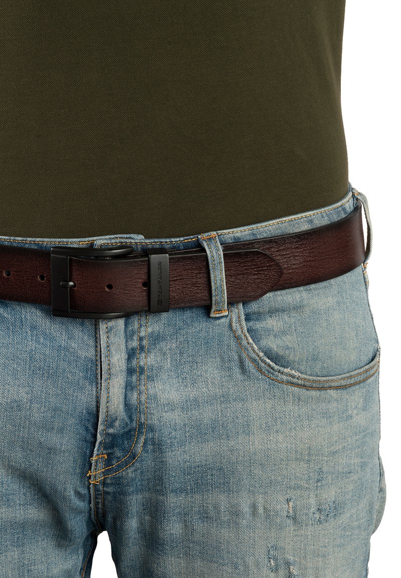 Bruine leren riem met een gestructureerde afwerking, voorzien van een matte zwarte metalen gesp. Draagt over vervaagde lichtblauwe spijkerbroek.