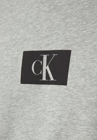 T-shirt in cotone grigio con un logo rettangolare nero con "cK" in carattere serif bianco, posizionato al centro in alto. Texture morbida.