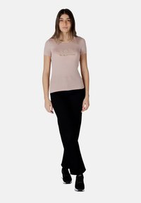 Camiseta ajustada de color rosa claro con mangas cortas, que presenta un brillante logotipo dorado "EA7 Emporio Armani". Combinada con pantalones negros de pierna recta y zapatillas negras.