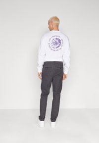 Camicetta bianca a maniche lunghe con grafica floreale viola sul retro, abbinata a pantaloni grigi. Scarpe bianche completano l'outfit.