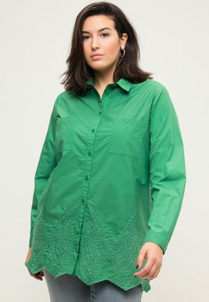 Une femme aux cheveux bruns porte une chemise verte à boutons avec un ourlet festonné brodé et un pantalon en jean bleu, se tenant devant un fond uni.
