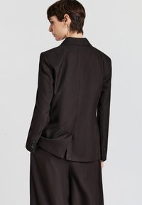 Blazer marrone su misura con una vestibilità strutturata, rever a lancia, spacco singolo sul retro e polsini con bottoni. Tessuto di trama liscia.