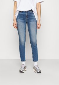 Persona con vaqueros azules deslavados, camiseta blanca, calcetines blancos y zapatillas plateadas de pie contra un fondo liso.