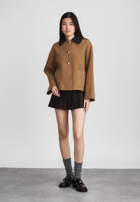 Vrouw in een oversized bruin jasje met zwarte kraag, een zwarte geplooide minirok, grijze sokken en zwarte instappers, staande voor een effen achtergrond.