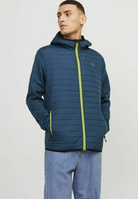 Giacca imbottita blu navy con cappuccio, caratterizzata da una cerniera e dettagli in verde lime a contrasto, tasche con zip e un design trapuntato.