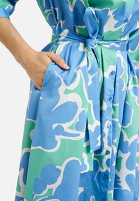 Blau-grün gemustertes Kleid mit gebundenem Bund, kurzen Ärmeln, Knöpfen auf der Vorderseite und Seitentaschen. Glattes Baumwollmaterial.