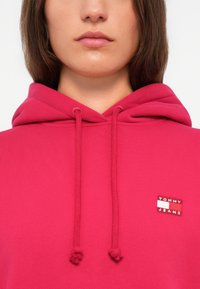 Rosa hettegenser laget av mykt stoff. Har snorstramming og et lite merketap med "TOMMY JEANS" i kontrastfarger.