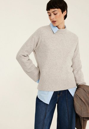 Femme aux cheveux courts et foncés portant un pull gris clair par-dessus une chemise bleue et un jean foncé, tenant une veste marron à carreaux dans une main.