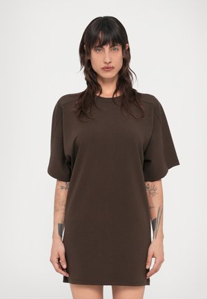 Žena s tmavými vlasmi v voľných tmavohnedých krátkych rukávoch, s viacerými tetovaniami na oboch predlakťiach, stojaca pred bielym pozadím.