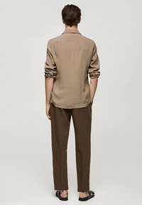 Mies, joka seisoo selkä kameraan päin, päällään vaaleanbeige pitkähihaisen paidan, jonka hihat on käännetty ylöspäin, ruskeat housut ja mustat sandaalit vaaleaa taustaa vasten.