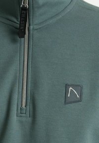 Pullover verde scuro con zip a metà lunghezza, realizzato in tessuto morbido. Presenta una piccola etichetta logo rettangolare nell'angolo in basso a destra.