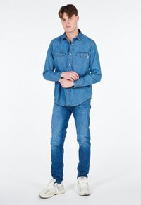 Camicia di jeans blu con un design classico, caratterizzata da due tasche sul petto e una chiusura a bottoni. Indossata con jeans blu abbinati e sneakers bianche.
