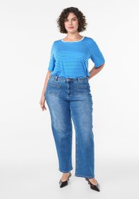 Žena s kučeravými vlasmi, oblečená v modro-bielom pruhovanom tričku, modrých džínsach s prednými vreckami a čiernych špicatých lodičkách, stojí s jednou rukou za chrbtom.