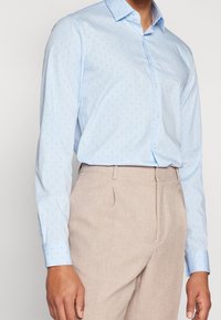 Světlomodrá košile na knoflíky s jemným vzorem, dlouhými rukávy a špičatým límcem, doplněná béžovými kalhotami.