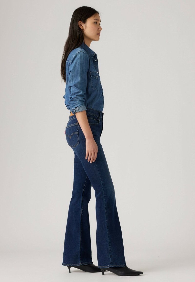 Femme debout de profil portant une chemise en jean, un jean évasé bleu foncé et des chaussures à talons noires sur un fond clair uni.