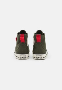 Un paio di sneaker alte Converse All Star verde scuro con linguette rosse sul tallone, esposte da dietro su uno sfondo bianco.