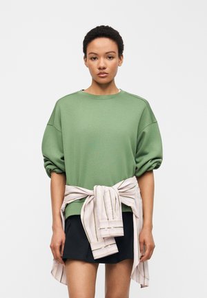 Woman with short curly hair wearing a loose green sweater, black skirt, and a striped shirt tied around her waist.