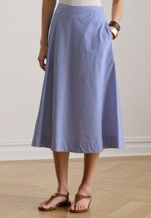 Kobieta ubraną w niebiesko-białą midi spódnicę w paski ukośne, biały top, brązowe sandały oraz masywne bransoletki na jednym nadgarstku, stojąca w pomieszczeniu.