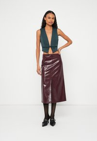 Frau steht vor weißem Hintergrund und trägt eine dunkelgrüne ärmellose Weste, die an der Taille gebunden ist, einen glänzenden burgunderroten Midirock, schwarze Strumpfhosen und spitze Schuhe.