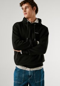 Μαύρη κουκούλα με κορδόνια, μπροστινή τσέπη και κεντημένο λογότυπο. Φορεμένο πάνω από ένα γκρίζο καρό πουκάμισο, συνδυασμένο με μπλε τζιν παντελόνι.