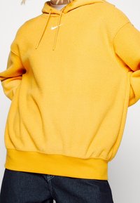 Keltainen fleece-huppari, jossa on kiristysnyörit hupussa, resorit hihansuissa ja helmassa sekä pieni valkoinen Nike-logo rinnassa.