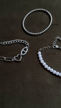 Három karkötő egy sötét felületen: egy ezüst lánc szívcsattal, egy ezüst gyöngyös, és egy áttetsző fehér gyöngyökkel.
