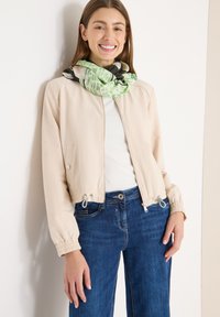 Giacca bomber corta beige con polsini elasticizzati, chiusura con zip e dettagli con coulisse. Indossata sopra una maglia bianca, abbinata a jeans blu e una sciarpa fantasia.