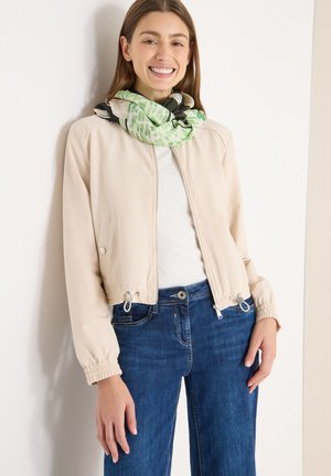 Beige cropped bomberjack met elastische boorden, ritssluiting en trekkoorddetails. Draagt het over een witte top, gecombineerd met blauwe spijkerbroek en een sjaal met patroon.