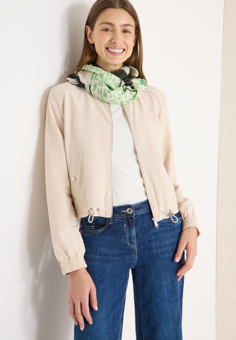 Giacca bomber corta beige con polsini elasticizzati, chiusura con zip e dettagli con coulisse. Indossata sopra una maglia bianca, abbinata a jeans blu e una sciarpa fantasia.