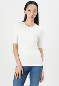 White ribbed short-sleeve t-shirt with a round neckline and small logo on the chest, paired with blue denim jeans. Smooth texture.