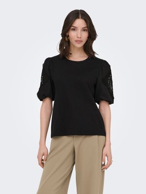 Kvinde iført sort bluse med pufærmer og blonde detaljer samt beige bukser med vide ben, stående mod en ensartet lys baggrund.