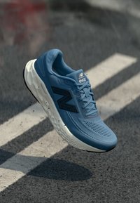 Modrá běžecká bota s texturovaným svrškem, prodyšným materiálem, měkkou bílou podrážkou a výrazným černým logem.