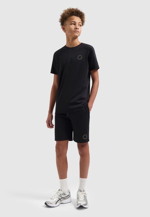 Tiener met zwart T-shirt en korte broek, witte sneakers en sokken, staat met één hand in de zak en kijkt opzij.
