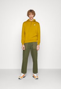 Sweatshirt amarelo com capuz e bolso frontal, combinado com calças largas verde-oliva e sapatilhas brancas com detalhes em laranja.