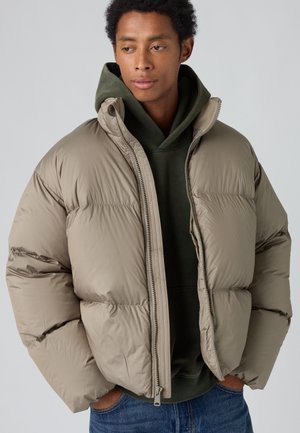 Jeune homme portant une doudoune marron clair par-dessus un sweat à capuche vert foncé, les mains dans les poches d’un jean bleu, regardant sur le côté.