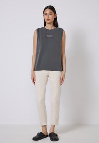 Mujer de pie en una blusa sin mangas de color gris oscuro con texto y pantalones beige, usando zapatos negros tipo slip-on, frente a un fondo claro y liso.