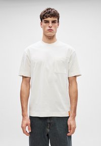 Λευκό βαμβακερό T-shirt με στρογγυλή λαιμόκοψη, κοντά μανίκια και τσέπη στο στήθος; διαθέτει μια πλευρική ετικέτα σε πορτοκαλί. Λείος υφασμάτινος χαρακτήρας, χαλαρή εφαρμογή.