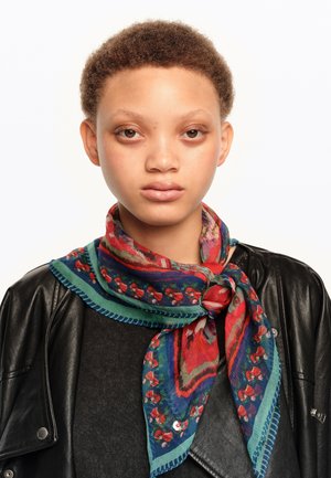 Persona joven con el cabello rizado y corto, que lleva una chaqueta de cuero negra y una bufanda colorida con estampado atada alrededor del cuello.