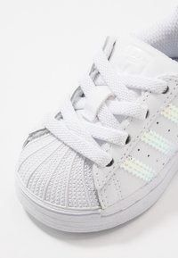 Vit sneakersko med texturerad tå i skalskydd, tre reflekterande ränder och mjuka skosnören. Tillverkad av läder och gummi med en slät yta.