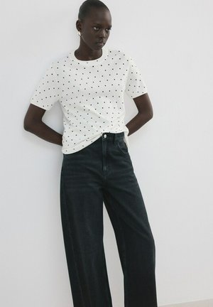 Femeie cu piele închisă la culoare, purtând un tricou alb cu mâneci scurte și buline negre și blugi negri largi, cu talie înaltă, stând cu mâinile la spate.