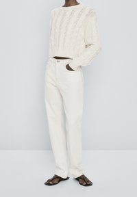 Pull en tricot crème à mailles, jeans blanc taille haute et tongs noires. La tenue présente un design texturé et une coupe décontractée.