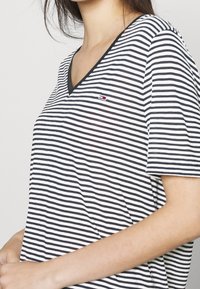 Žena v černobíle pruhovaném V-krku tričku s krátkými rukávy a malým logem na hrudi.